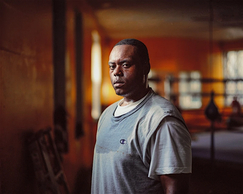 Portrait of an African-American man in a blue Champion shirt, looking at the camera in an orange-toned indoor gym setting.