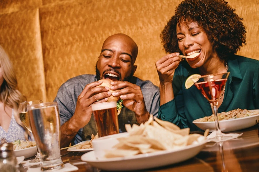 A man and woman at a restaurant, enjoying burgers, fries, and cocktails, with a blonde woman visible in the background in a warm setting.