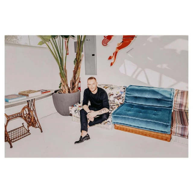 Stylish adult man with ginger hair sits on a patterned couch in a bright room, featuring a large potted plant and artistic wall decor.
