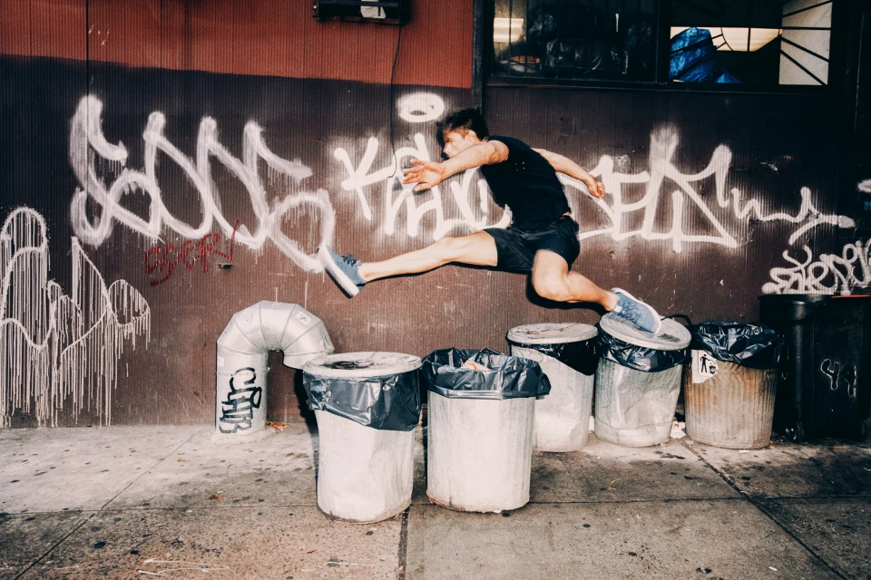 A man in mid-air leaps over trash cans in an urban setting with a graffiti-covered wall, showcasing a dynamic action shot in daylight.