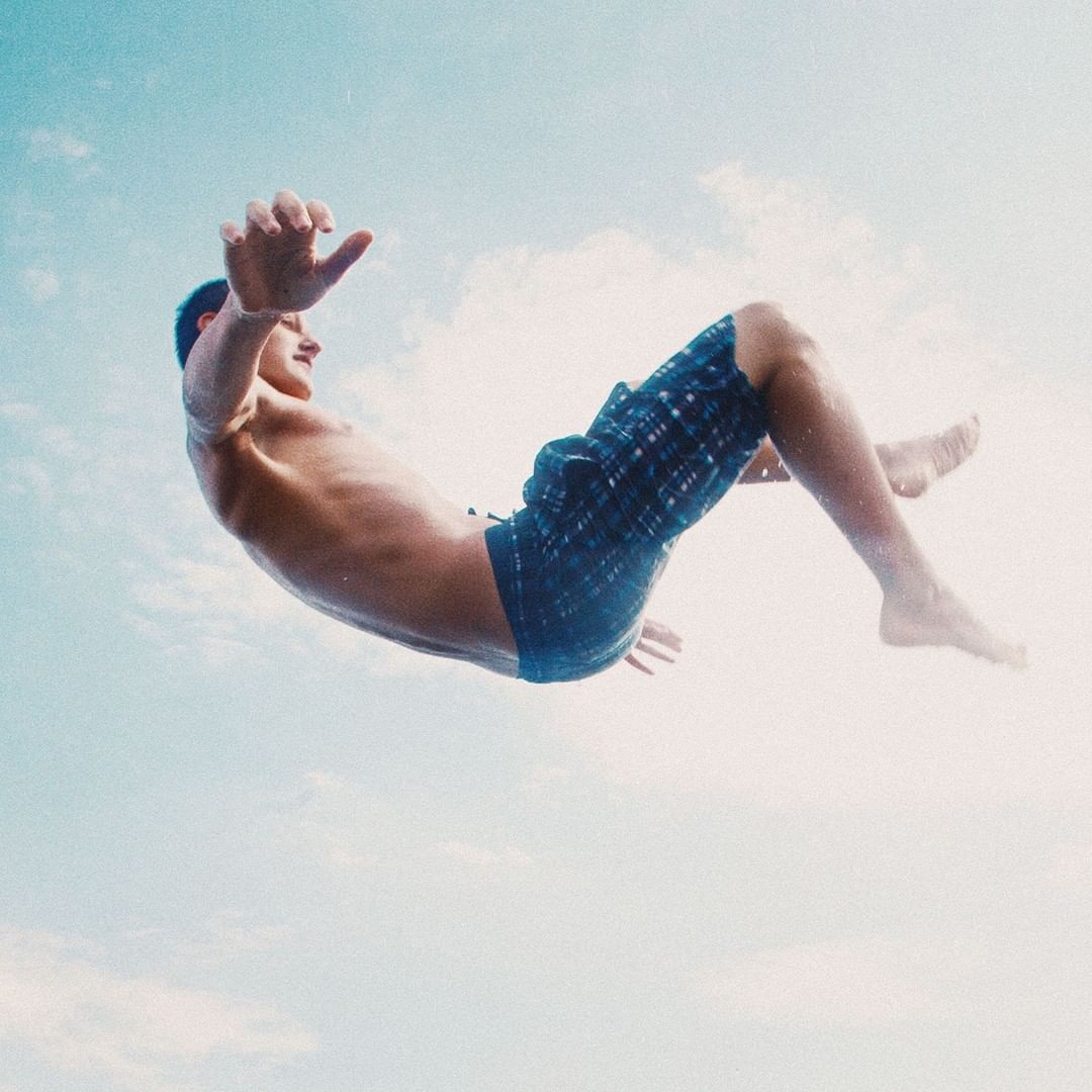 Young man in mid-air, wearing blue patterned swim trunks, seen from below against a soft blue sky with faint white clouds.