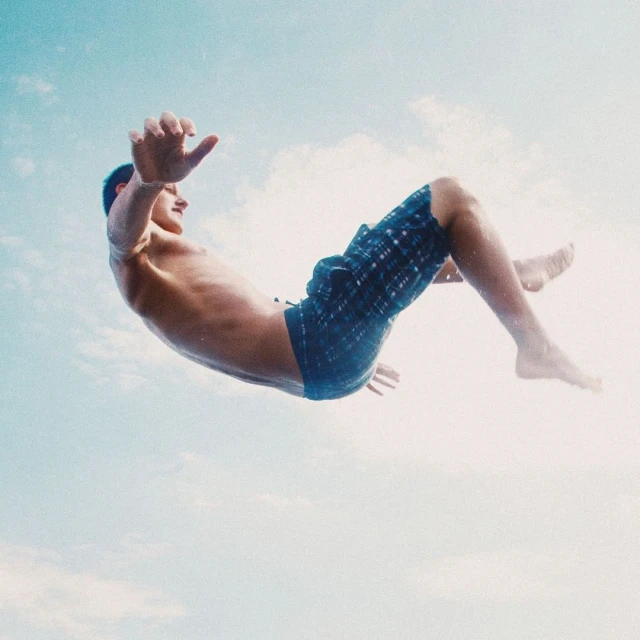 Young man in mid-air, wearing blue patterned swim trunks, seen from below against a soft blue sky with faint white clouds.