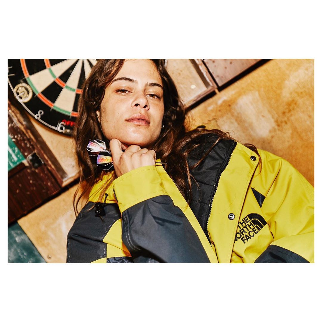 A young woman in a yellow and black jacket poses indoors with a dartboard visible. The scene features a gritty, urban setting.