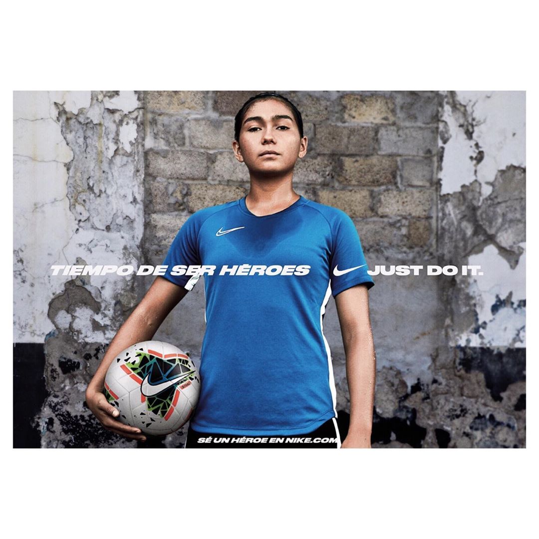 A young adult female athlete holds a soccer ball, wearing a blue Nike soccer jersey, photographed against a textured grey brick wall.
