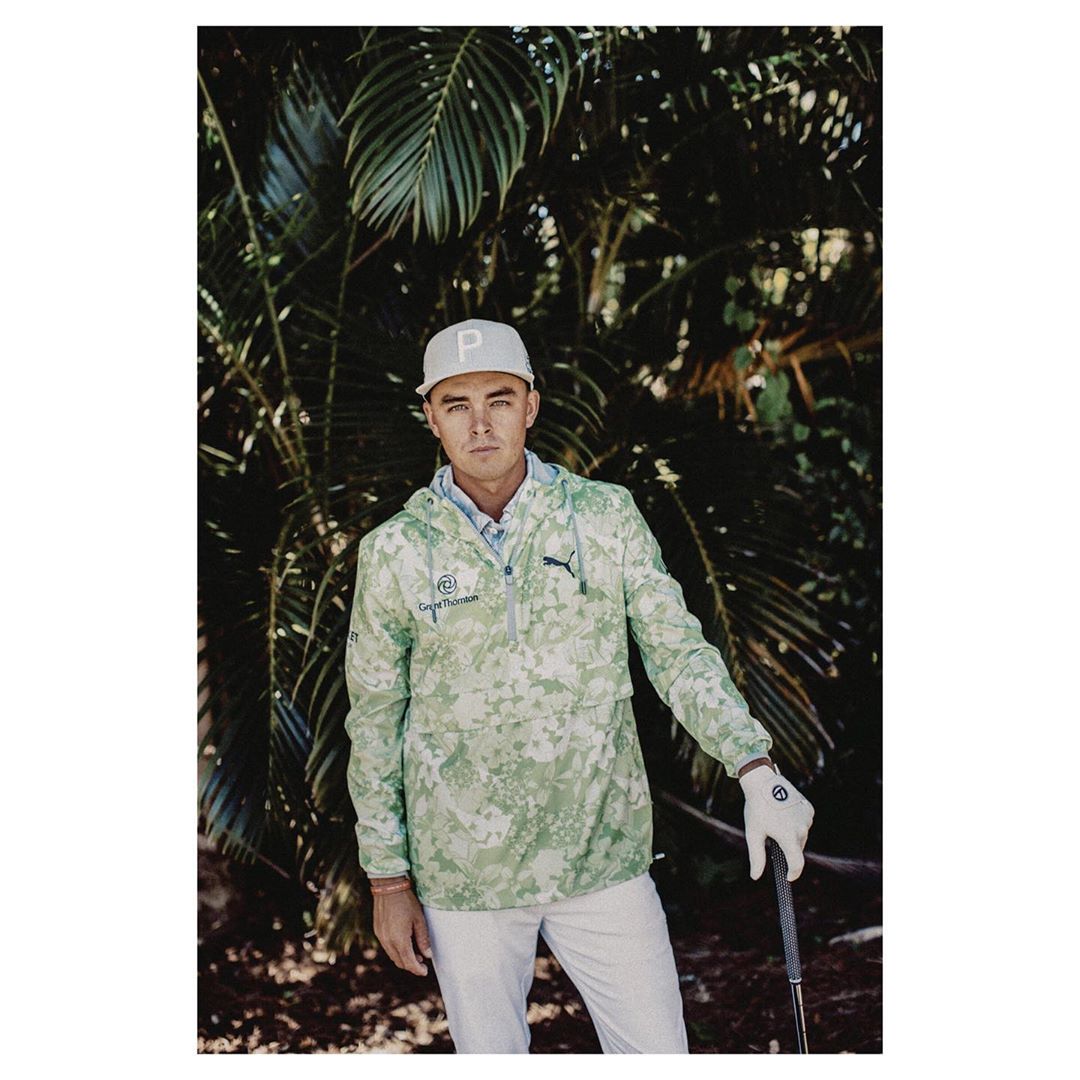 Male model in a camouflage jacket and white pants poses outdoors, wearing a baseball cap and golf glove in a lush tropical setting.