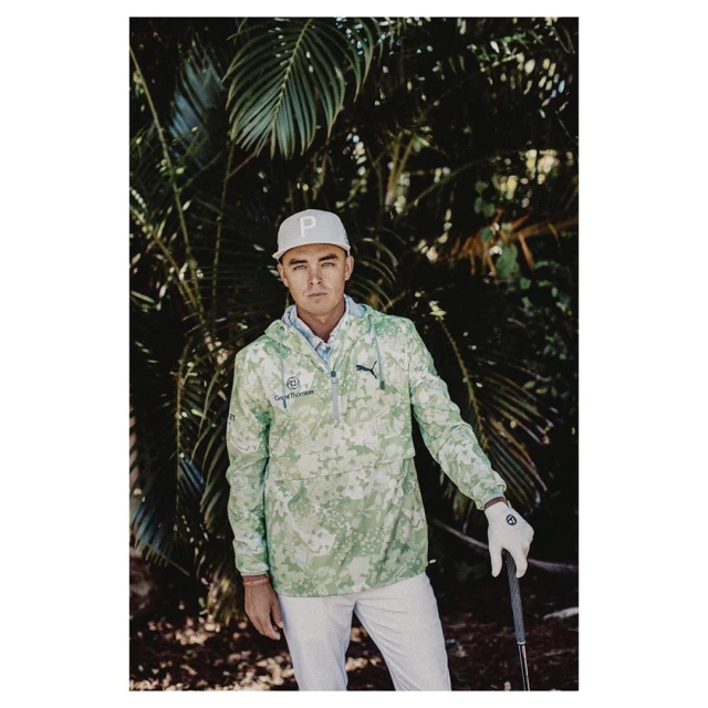 Male model in a camouflage jacket and white pants poses outdoors, wearing a baseball cap and golf glove in a lush tropical setting.