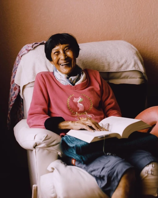 Elderly Asian woman smiles while sitting in a white armchair, holding a book, wearing a pink sweatshirt in an indoor setting.