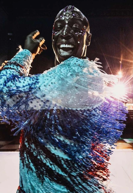Smiling adult wearing a decorated costume with blue and white fringe stands on stage lit with bright spotlights in a performance venue.