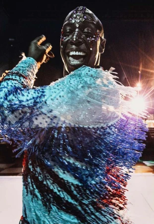 Smiling adult wearing a decorated costume with blue and white fringe stands on stage lit with bright spotlights in a performance venue.