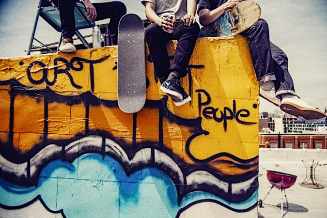 Three people sit on a rooftop wall next to graffiti with skateboards, an outdoor grill, and buildings in the background on a sunny day.