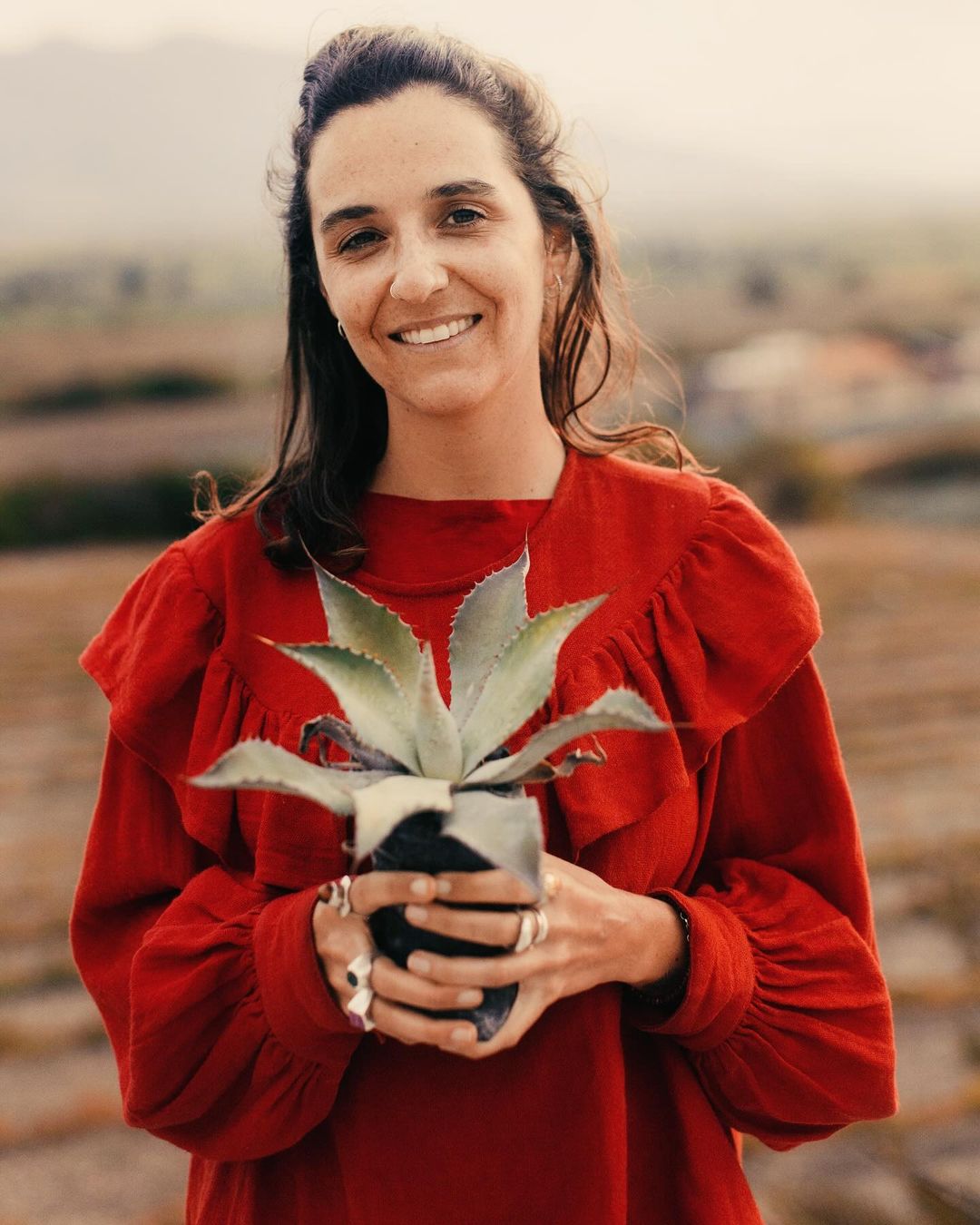 Woman in red dress outdoors holding an agave plant in a pot. Background shows an arid landscape with rows of low-lying plants.