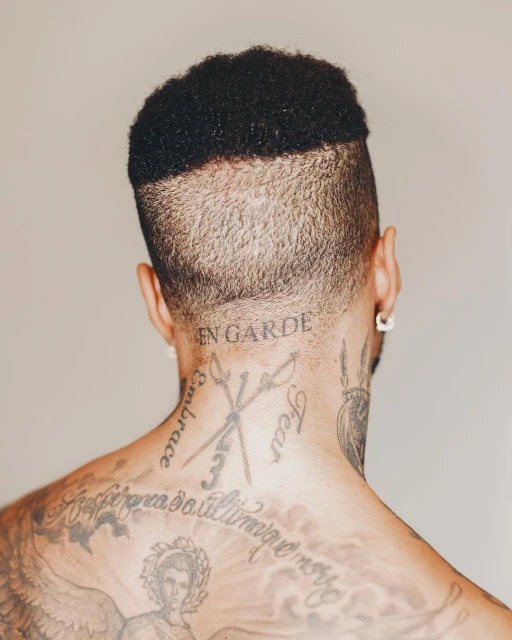 Back view of a person with short dark hair and tattoos covering the back and shoulders against a neutral background, showcasing body art.