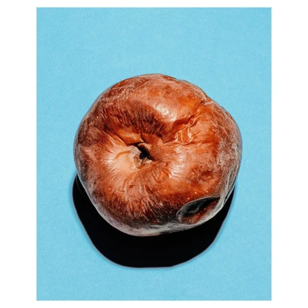Close-up of a decaying apple with a bruised skin texture and dark spots against a bright blue background, casting a subtle shadow.