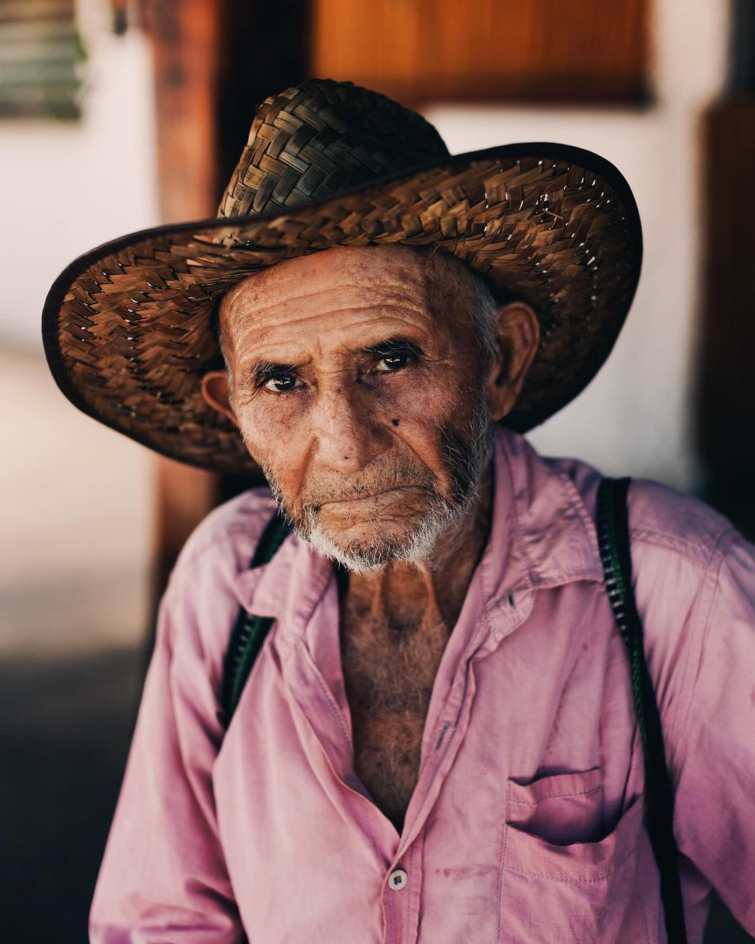 Close-up portrait of an elderly man with a straw hat, wearing a pink collared shirt with suspenders in an outdoor setting.