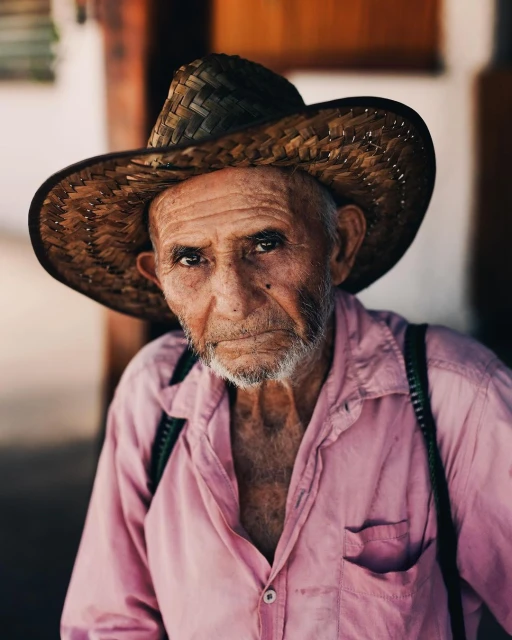 Close-up portrait of an elderly man with a straw hat, wearing a pink collared shirt with suspenders in an outdoor setting.
