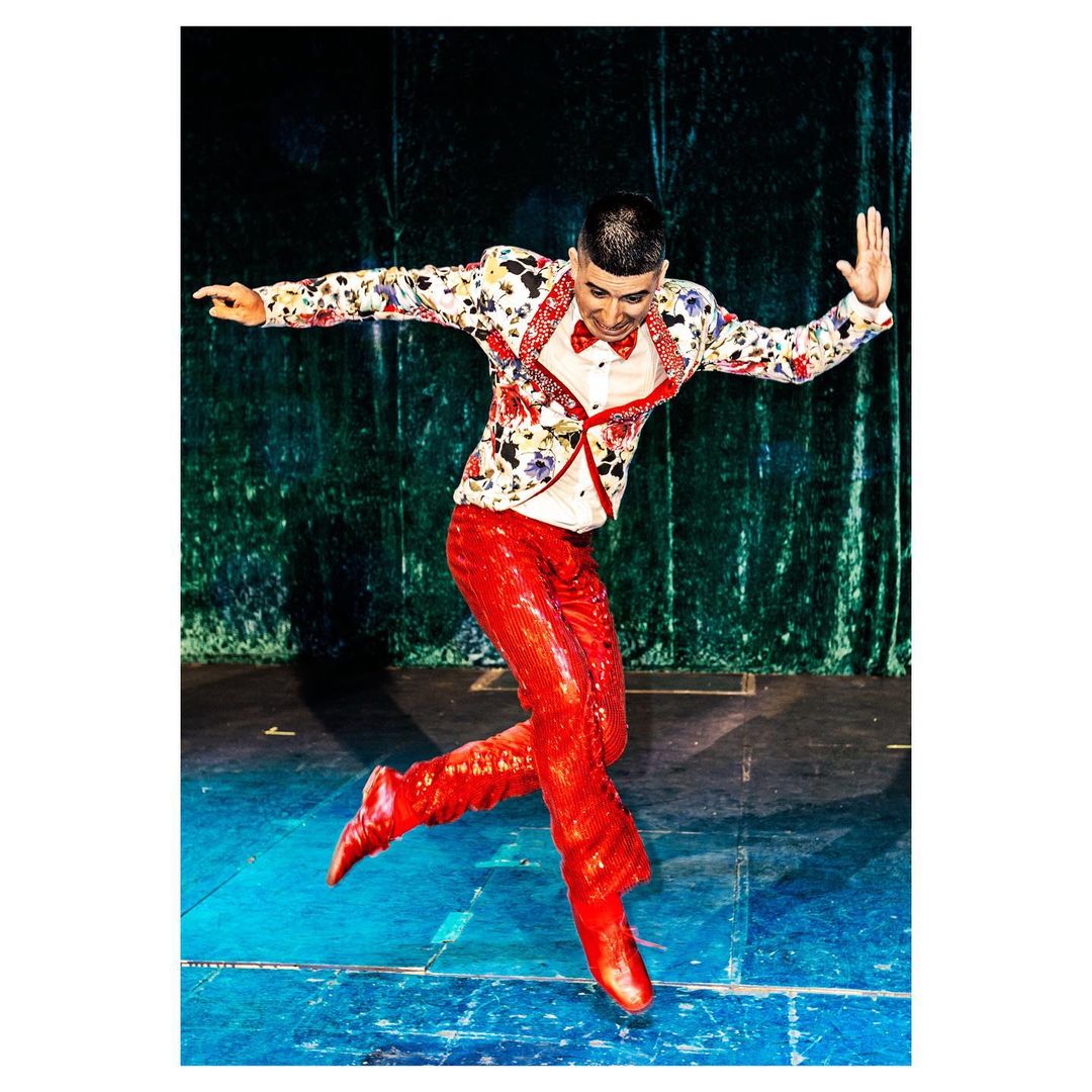 A male dancer in a floral jacket and shiny red pants leaps on stage with a green velvet curtain backdrop in a vibrant performance shot.