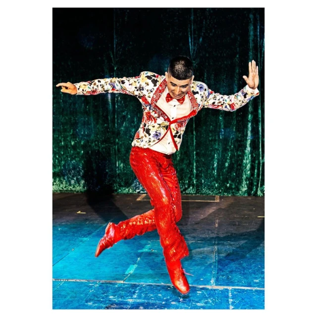 A male dancer in a floral jacket and shiny red pants leaps on stage with a green velvet curtain backdrop in a vibrant performance shot.