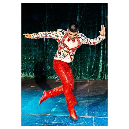 A male dancer in a floral jacket and shiny red pants leaps on stage with a green velvet curtain backdrop in a vibrant performance shot.