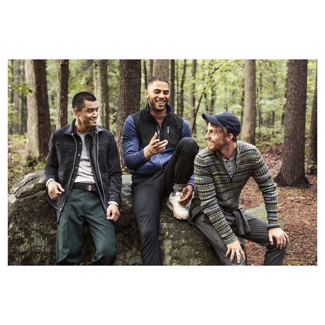 Three adult men are sitting on a large rock in a forest setting, smiling and talking. They are dressed in casual, outdoor clothing.