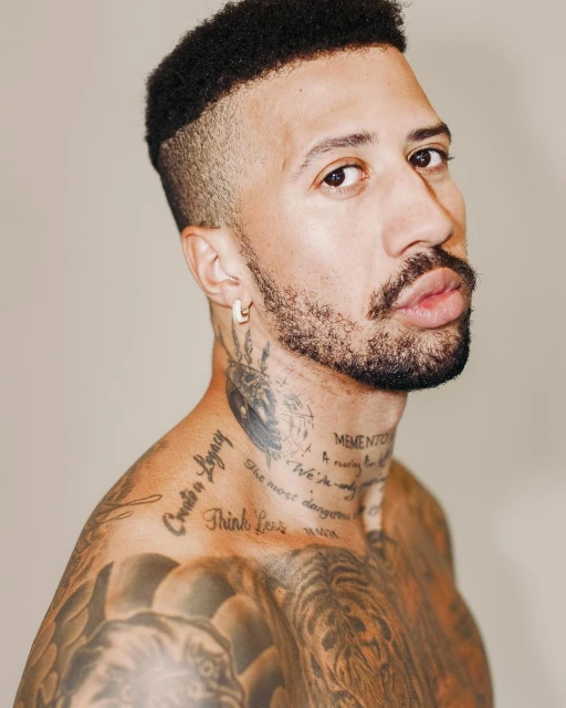 Close-up studio portrait of a tattooed adult male with a trimmed afro, gold earring, and a neutral facial expression against a neutral background.