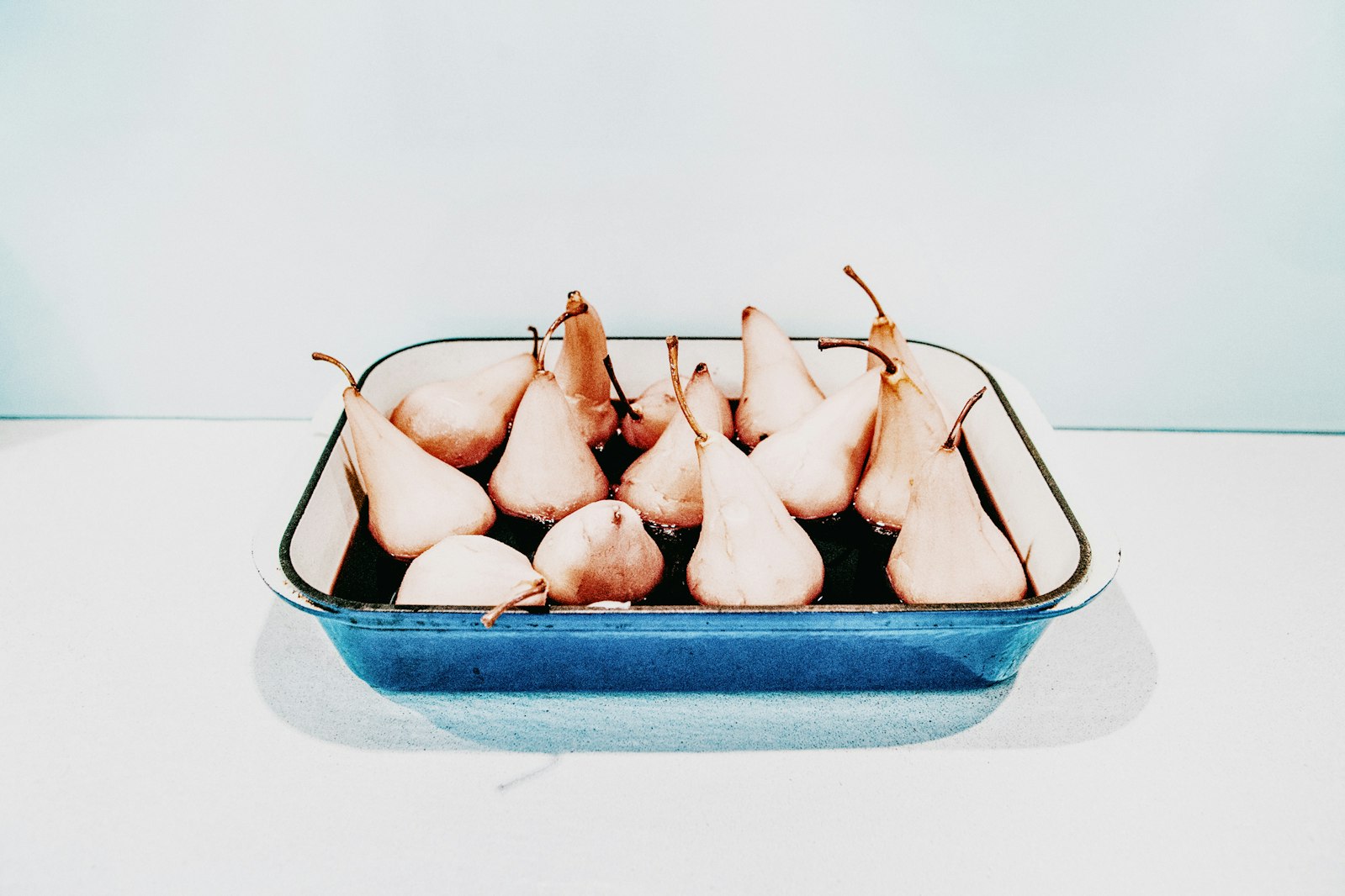 Poached pears in a blue baking dish against a white and light blue background, studio lighting, minimalist food still life.