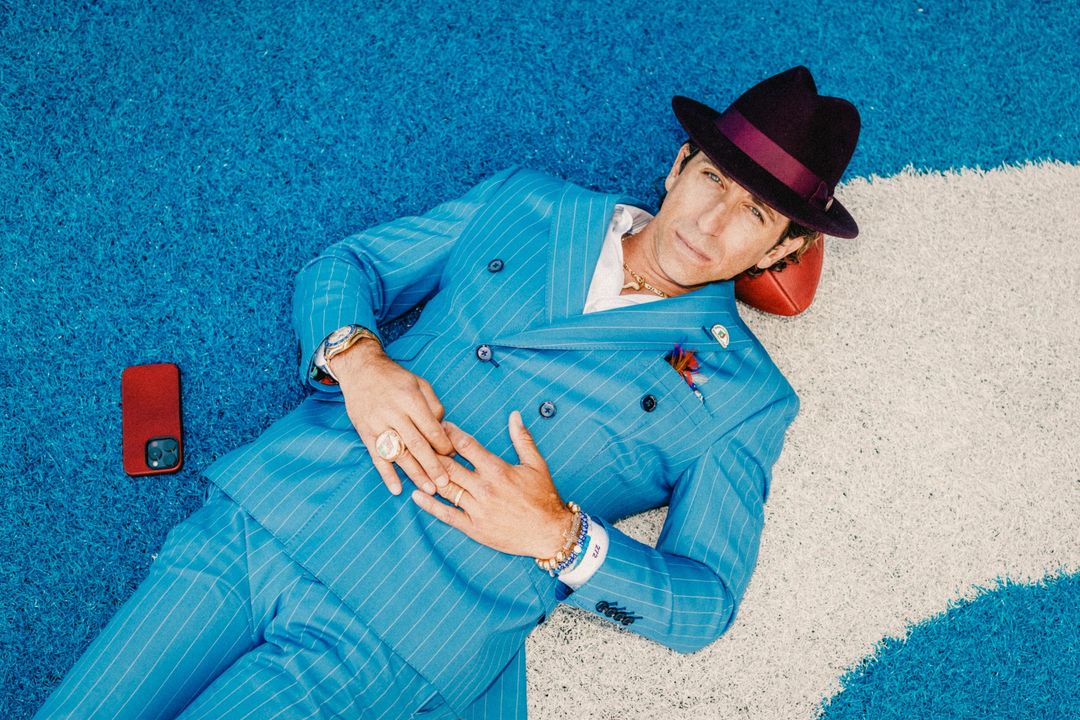 A stylish man in a blue pinstripe suit and fedora lies on a blue turf, resting his head on a football. He wears jewelry, and a phone is nearby.