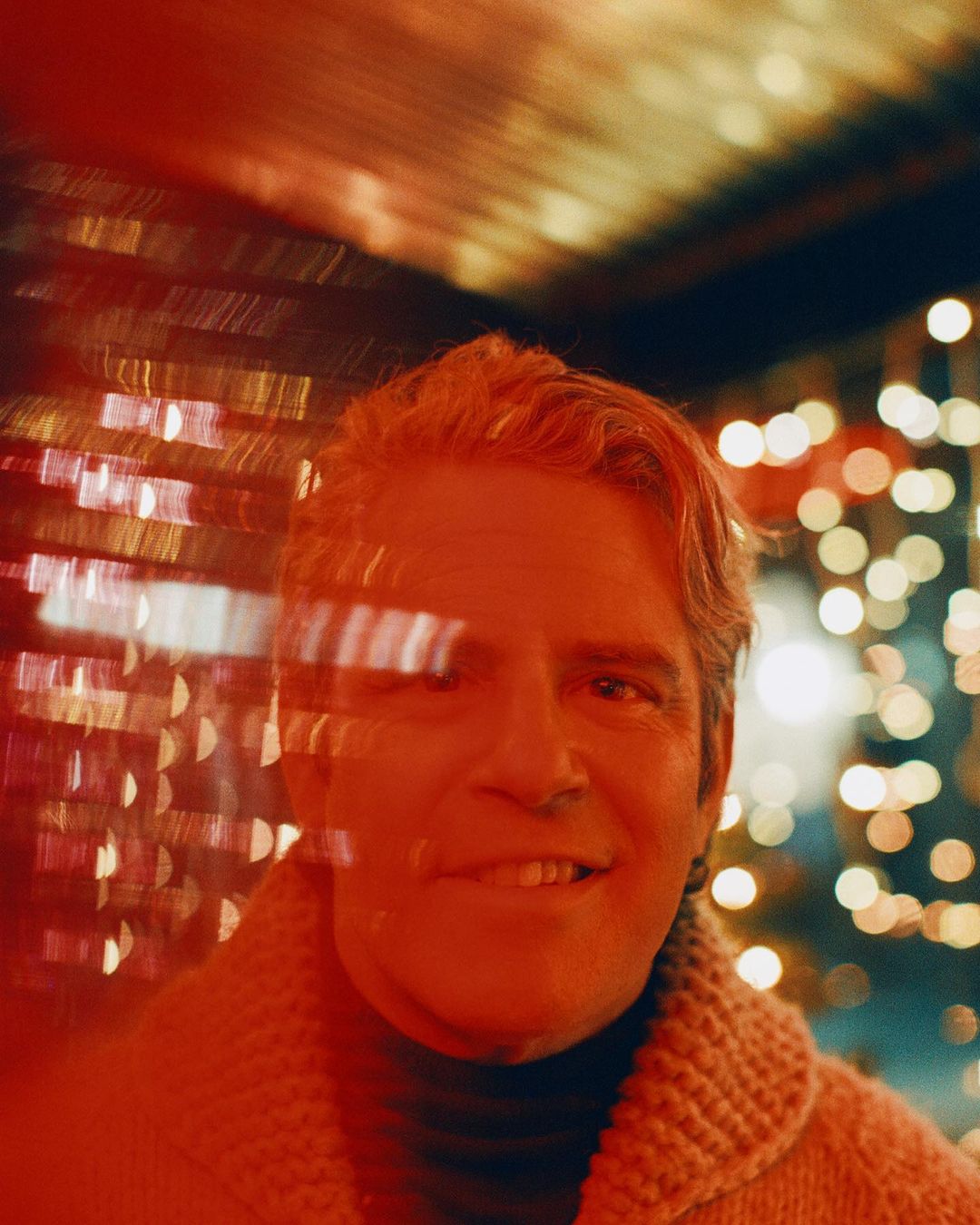 Close-up portrait of a smiling adult male with grey hair wearing a knitted cardigan, against a bokeh light background.