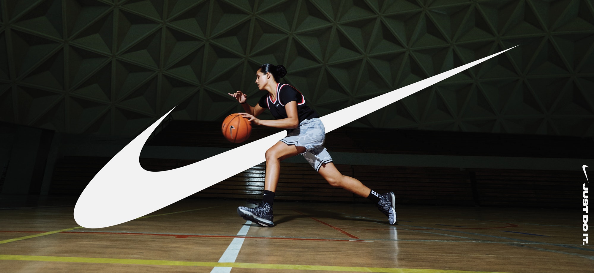 A female basketball player dribbles a basketball in an indoor court with a prominent Nike logo overlaid on the image.