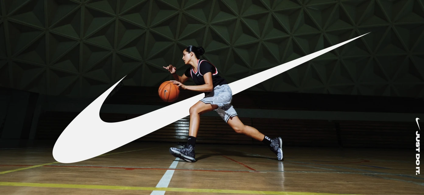 A female basketball player dribbles a basketball in an indoor court with a prominent Nike logo overlaid on the image.