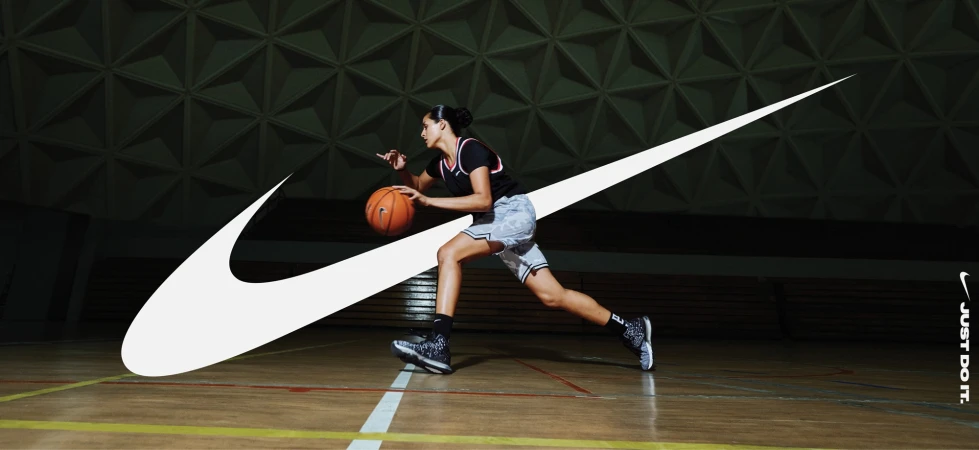 A female basketball player dribbles a basketball in an indoor court with a prominent Nike logo overlaid on the image.