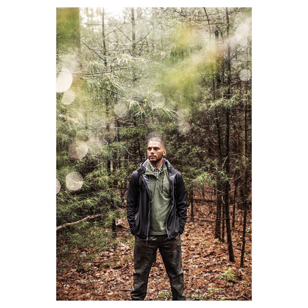 A man stands confidently in a forest wearing a black jacket and pants. Pine trees surround him, creating a natural outdoor setting.