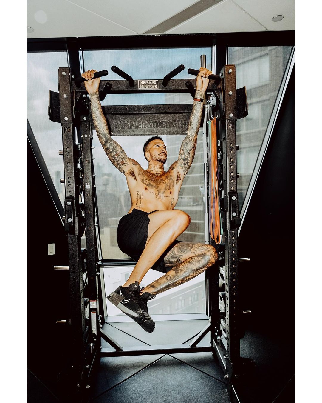 Tattooed man in black shorts executes a pull-up in a home gym on a Hammer Strength rack, exhibiting muscular physique and fitness.