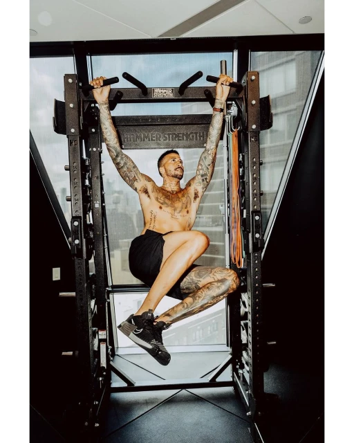 Tattooed man in black shorts executes a pull-up in a home gym on a Hammer Strength rack, exhibiting muscular physique and fitness.