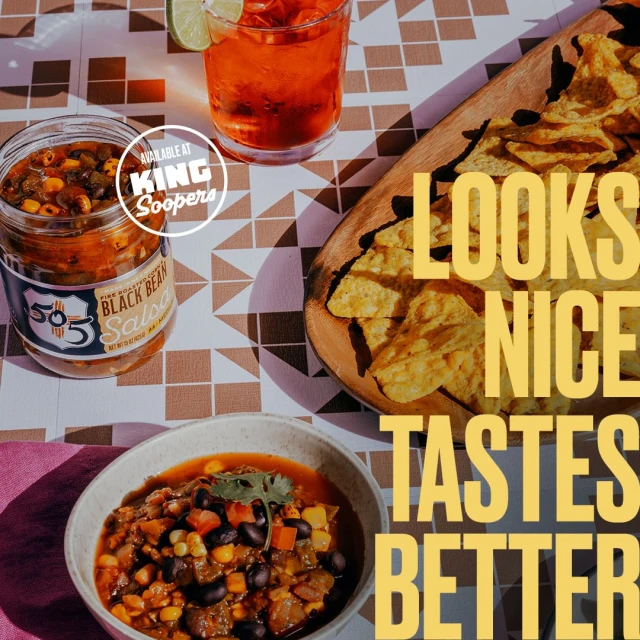 Overhead shot of a jar of black bean salsa, tortilla chips on a wooden board, and a drink with a lime wedge on a patterned surface.