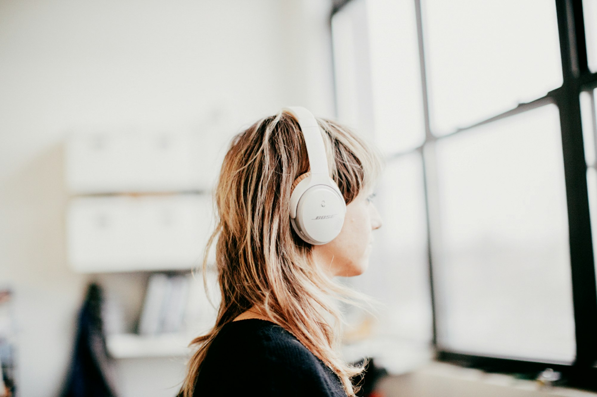 Woman with blonde hair wearing white headphones looks out window in bright interior setting with natural light. Music and audio concept.