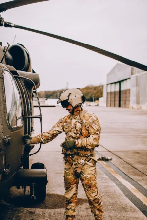 Military personnel in camouflage uniform stands next to a parked helicopter; neutral expression, natural daylight, outdoor setting.