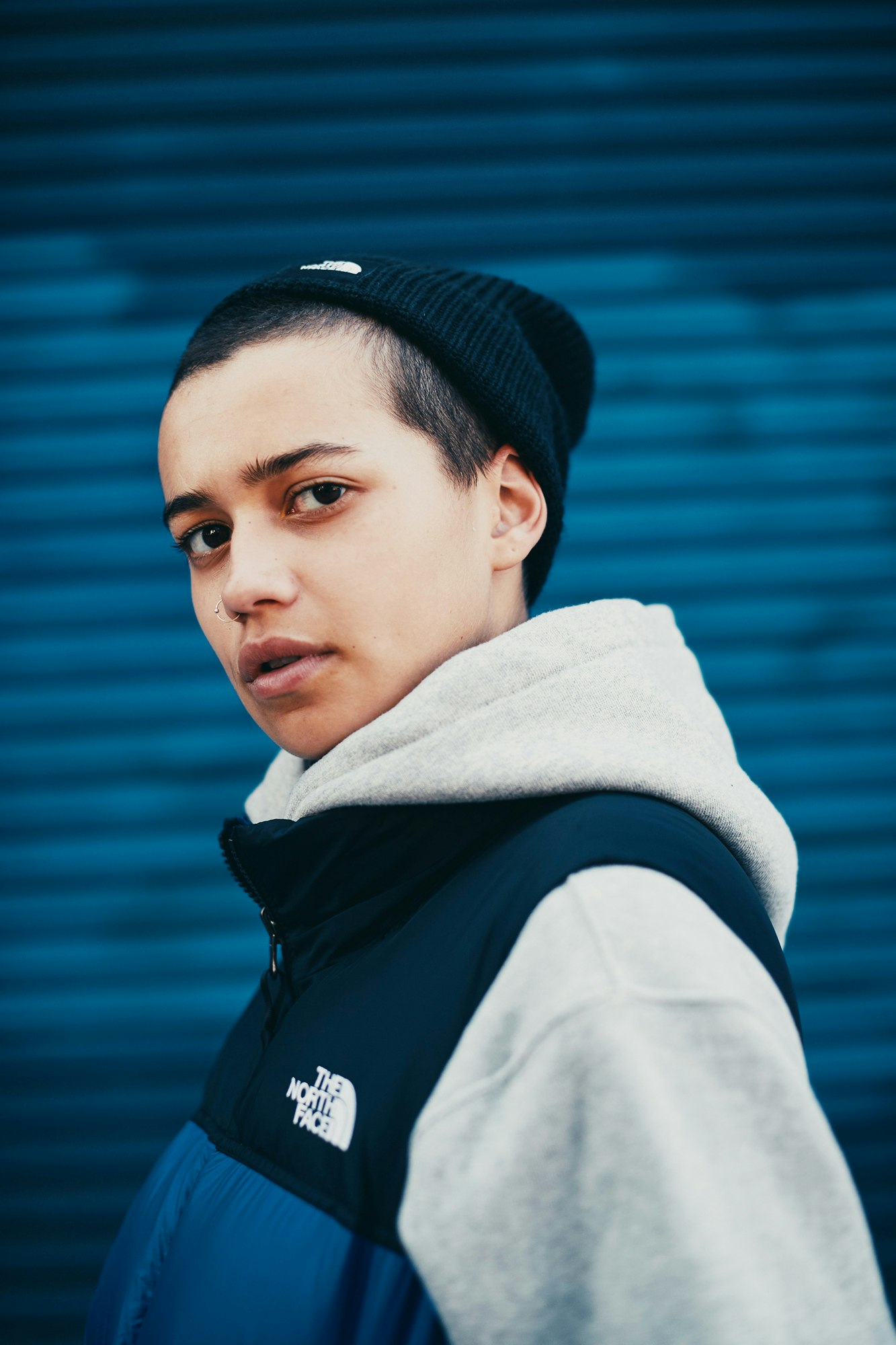 Close-up portrait of a young adult wearing a beanie and a gray hooded sweatshirt layered under a blue and black jacket.