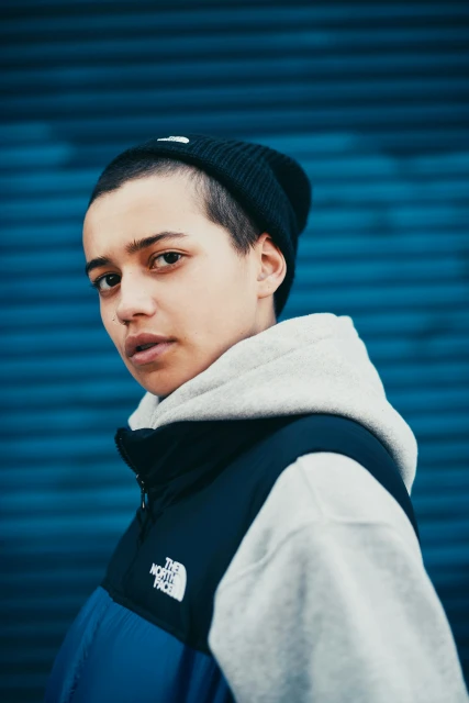 Close-up portrait of a young adult wearing a beanie and a gray hooded sweatshirt layered under a blue and black jacket.