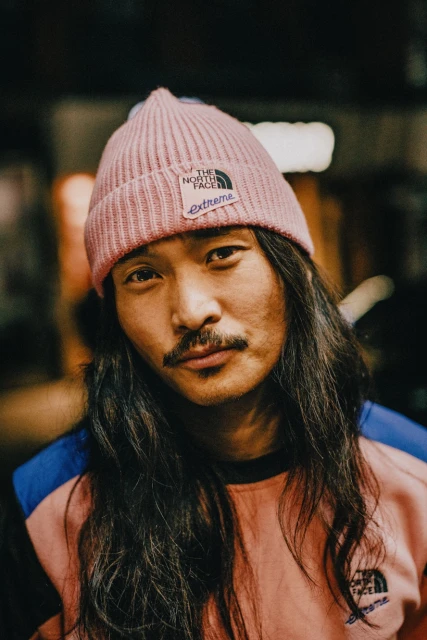 Portrait of an adult male wearing a pink beanie with long dark hair, a mustache, and a colorful sweatshirt, looking directly at the camera.