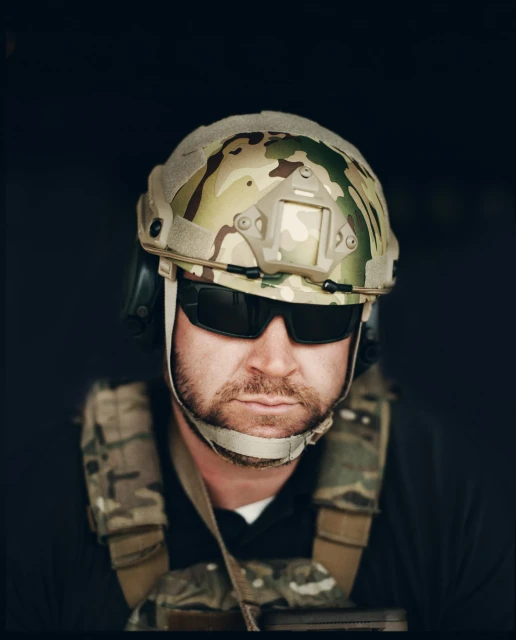 Close-up of a serious adult male with fair skin wearing a camo helmet and sunglasses, with tactical gear against a dark backdrop.