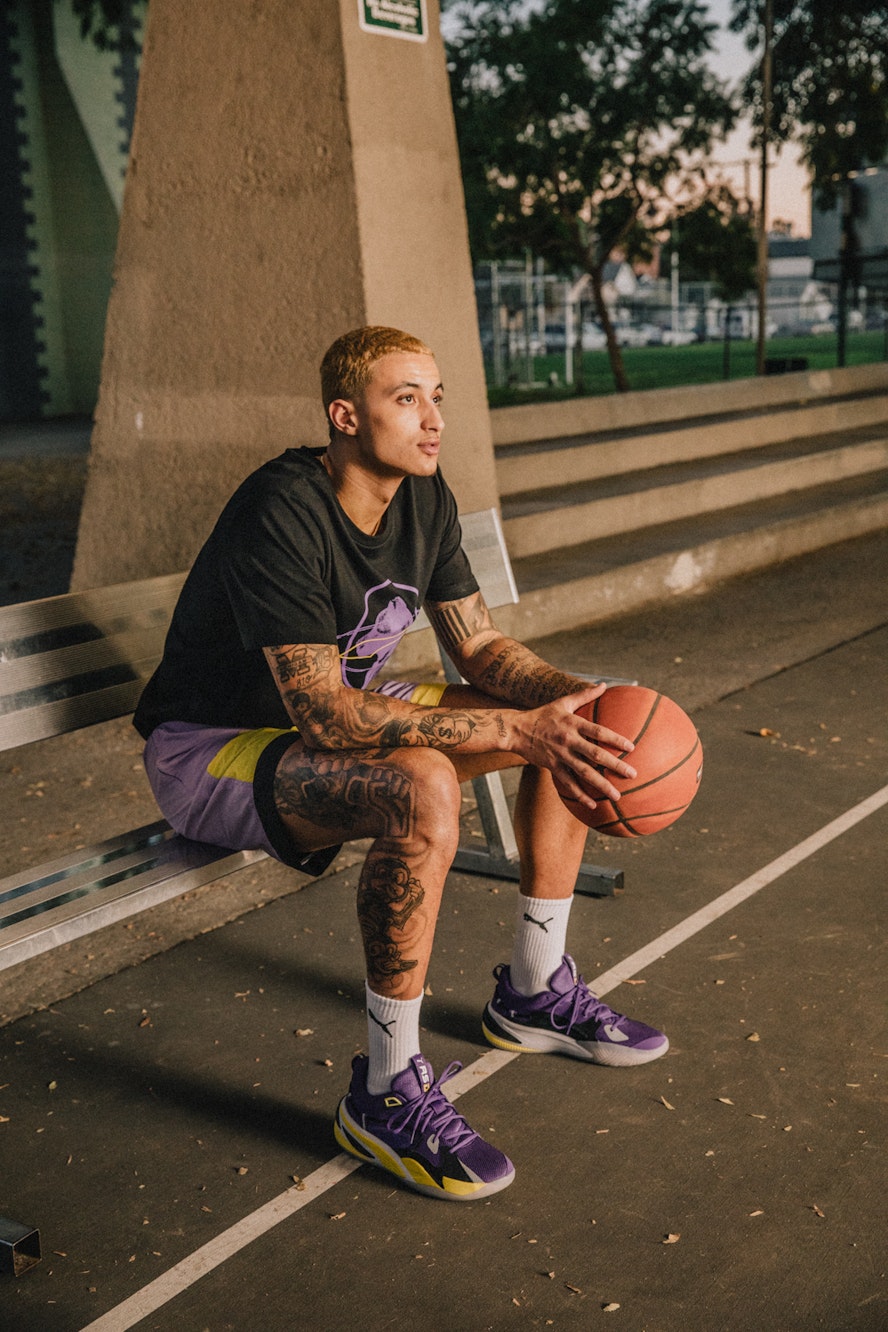 An athletic man sits on an outdoor bench holding a basketball while wearing branded PUMA athletic shoes and a black graphic tee shirt.