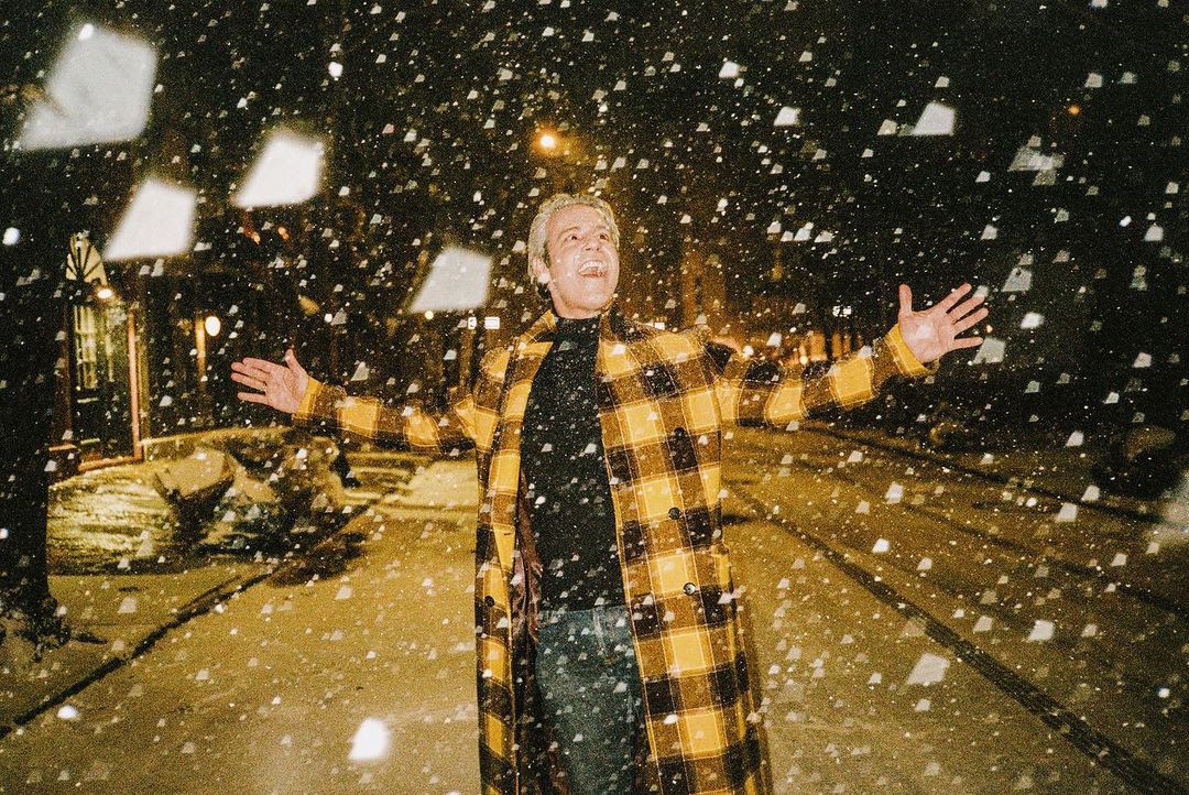 Adult with arms open outside at night, under falling snow. He wears a yellow plaid coat, black shirt, and blue jeans.