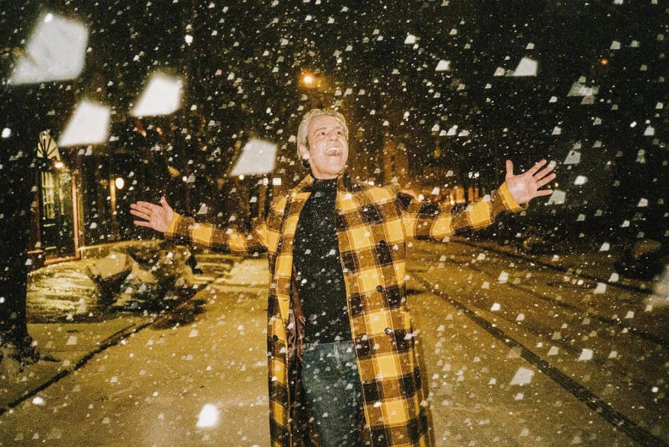 Adult with arms open outside at night, under falling snow. He wears a yellow plaid coat, black shirt, and blue jeans.