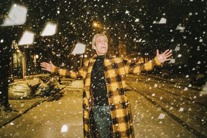 Adult with arms open outside at night, under falling snow. He wears a yellow plaid coat, black shirt, and blue jeans.