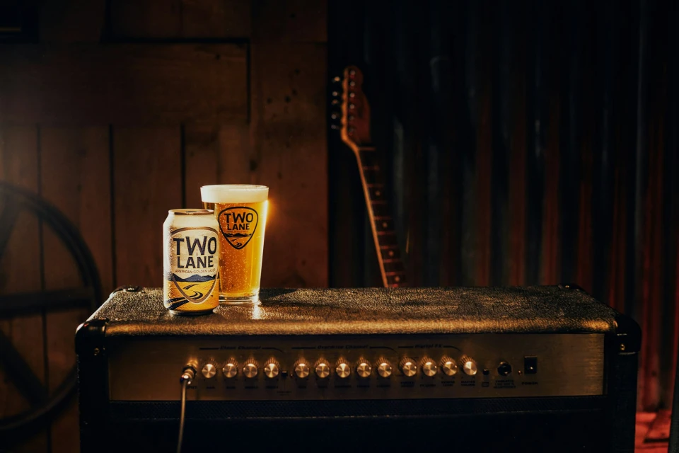 A can and glass of Two Lane American Golden Lager sit atop a guitar amplifier in a dim music venue with a rustic vibe.