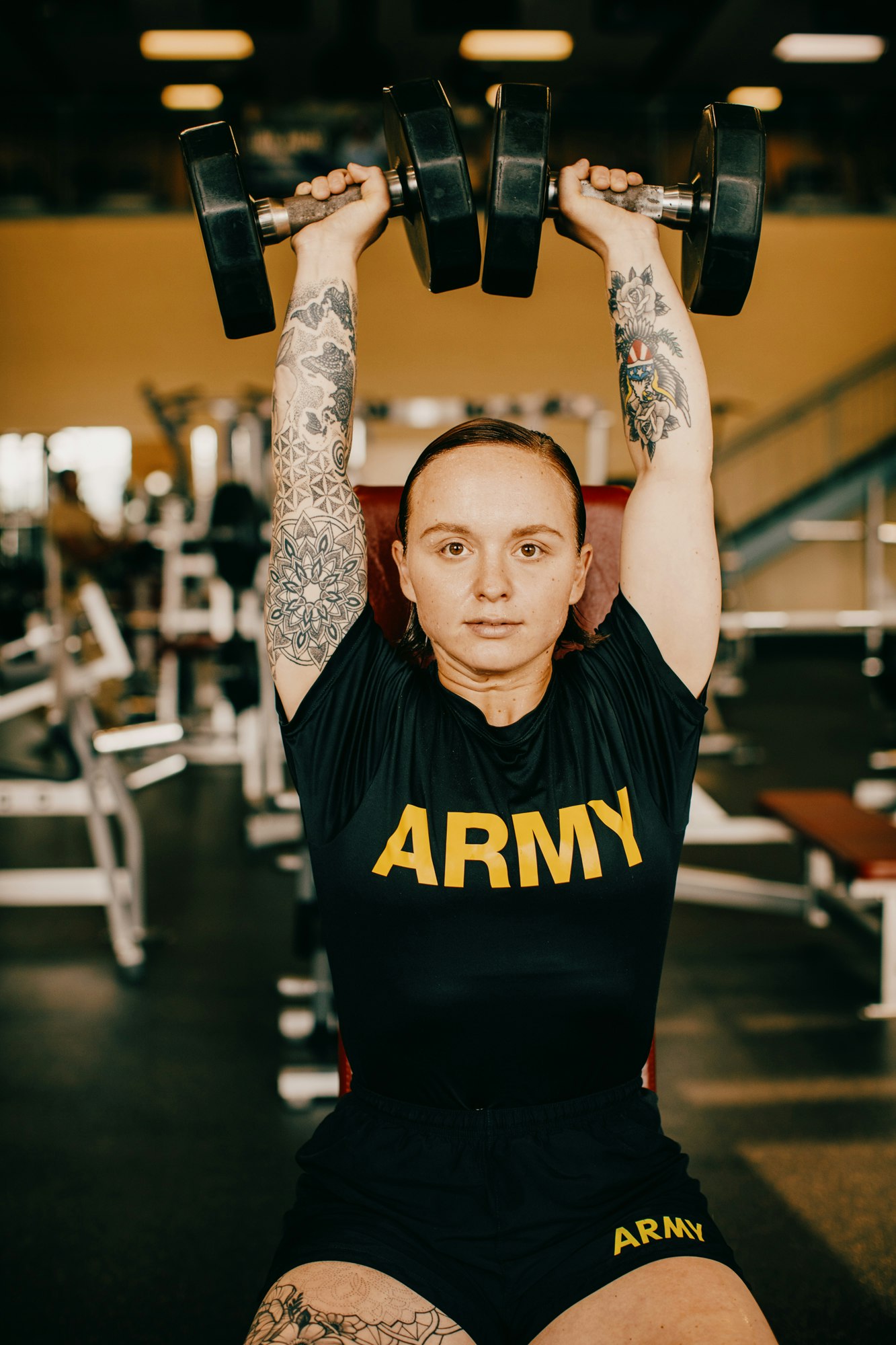 A woman in workout clothing lifts dumbbells overhead at a gym, displaying arm tattoos, set against gym equipment and an orange wall.