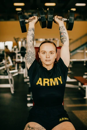 A woman in workout clothing lifts dumbbells overhead at a gym, displaying arm tattoos, set against gym equipment and an orange wall.