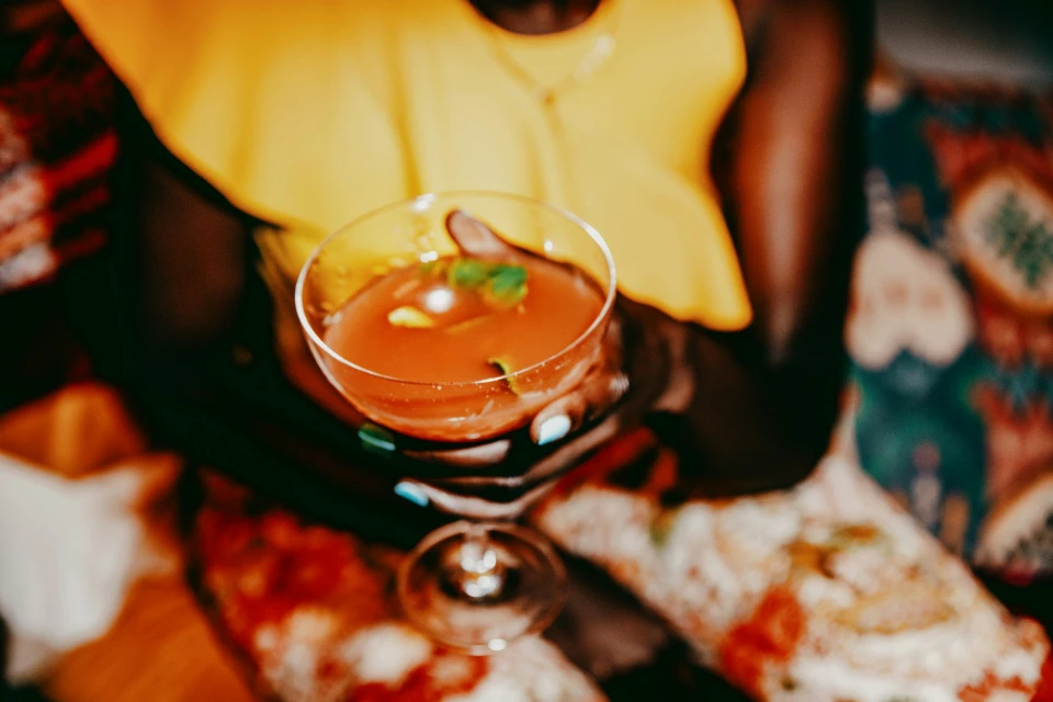 Person holding a glass of red cocktail with mint garnish, wearing a yellow top, against a patterned textile background.