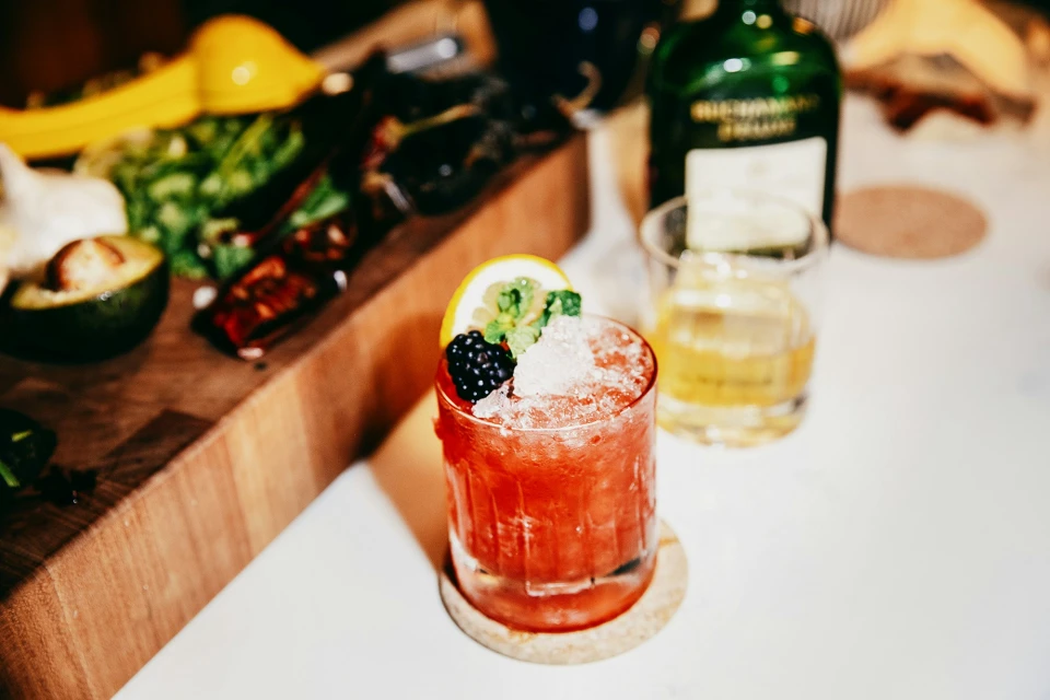 Red cocktail in glass with lemon, blackberry and crushed ice, alongside avocado, lime, and bottle on wooden cutting board.
