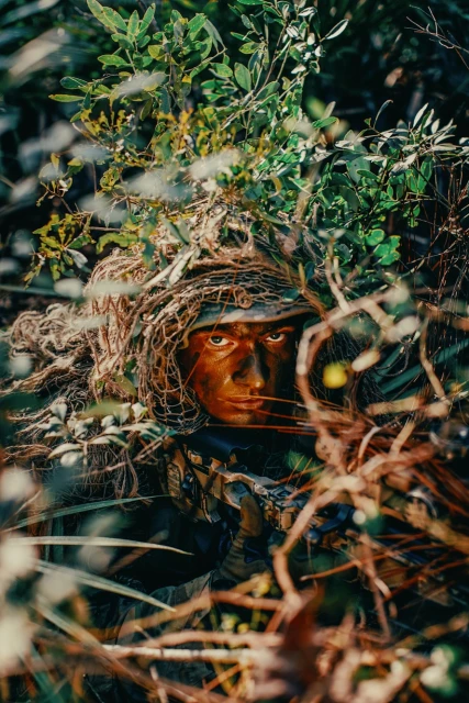 Crouched adult male wearing camouflage and face paint hides in a densely overgrown environment, appearing to be a soldier or hunter.
